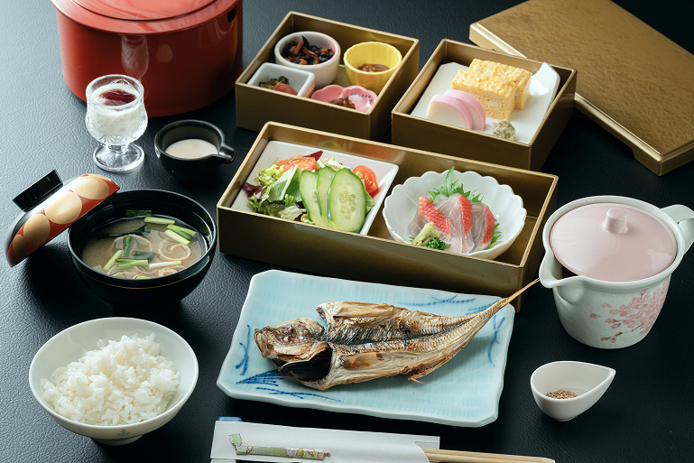 朝食膳。焼き魚、ご飯、味噌汁、お重のおかず(サラダ、蒲鉾、卵焼き、香の物)、とろろ芋、温泉卵、飲み物、急須、箸が黒いテーブルに並ぶ、彩り豊かで滋味深い和食。