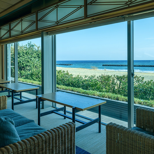 大きな窓から青い海と砂浜が見渡せる、明るい休憩スペース。手前には籐製のソファとテーブルがあり、奥にもテーブルと椅子が配置されている