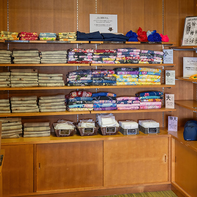 客室用の浴衣コーナー。棚にはサイズ別に分けられた色とりどりの浴衣が整然と並べられ、小・中・大・特大の表示がある。下段には帯や小物が入ったかごが置かれている。