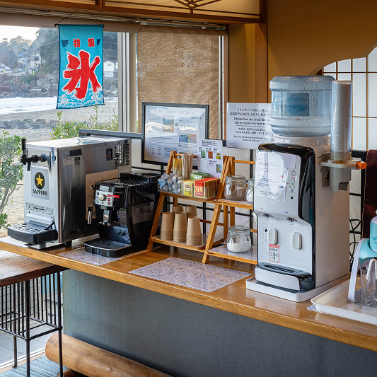 ドリンクコーナー。左からビールサーバー、コーヒーマシン、紙コップと紅茶などが並ぶ棚、ウォーターサーバーが設置されている。窓の外には川辺の風景が広がり、氷の旗が吊るされている。