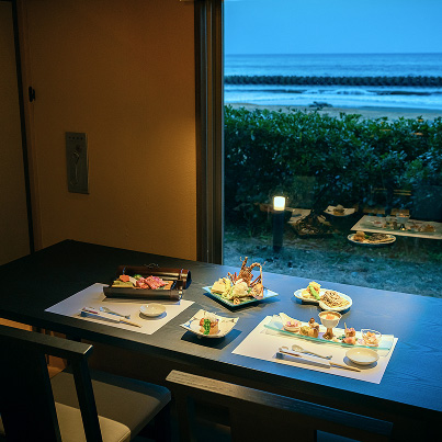 夜の海辺を望む窓際のテーブルに、複数の上品な料理が並ぶ様子。