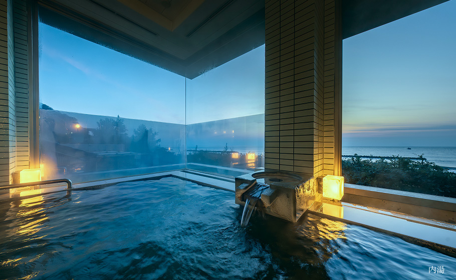 夕暮れ時の海を一望できる、大きな窓に囲まれた屋内温泉。湯口からお湯が流れ、穏やかな雰囲気が広がっています。