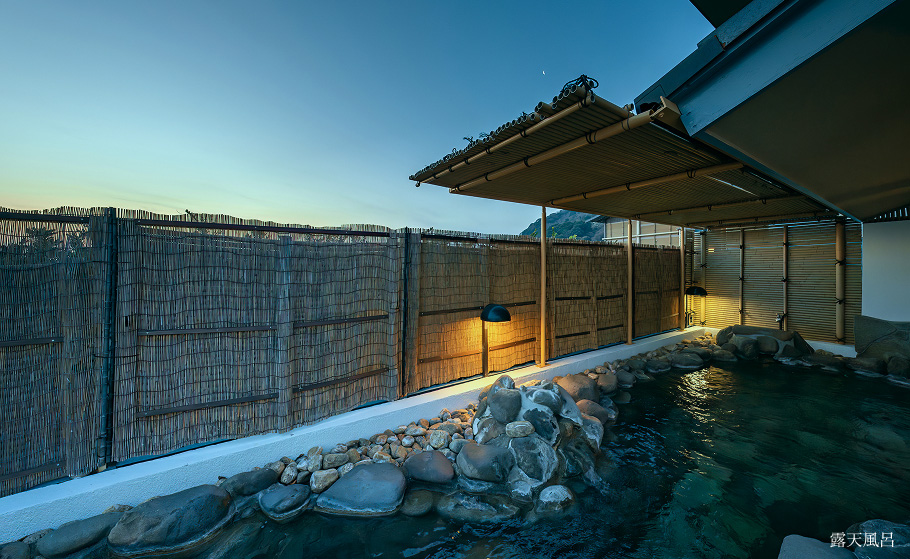 岩に囲まれた和風の露天風呂。夕暮れ空の下、竹垣と照明に包まれた静かな湯船が広がります。