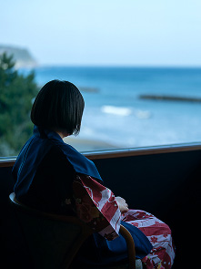 女性は椅子に座り、窓の外の穏やかな海を見つめている。水平線はぼやけており、空と海の色が溶け合っているように見える。