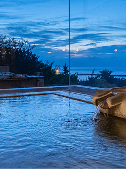 夜明け前の露天風呂の様子。青みがかった暗い空には、雲が広がり、水平線付近は明るくなり始めている。湯船の水は静かに波打ち、湯口からはお湯が流れ込んでいる。