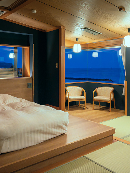 夜の海が見える、和風のベッドルーム。手前には、低い木製の台に布団が敷かれたベッドがあり、白いストライプの寝具がかかっている。ベッドの奥には、大きな窓があり、濃い青色の海が映っている。