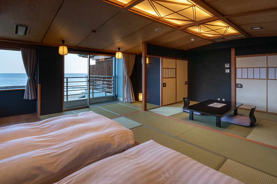 海辺の和室の広々とした空間。手前には、淡いピンクと白のストライプ柄のシーツがかけられた二つのベッドが並んでいる。ベッドの奥は畳敷きのスペースで、中央には黒いローテーブルと座椅子が二つ置かれている。