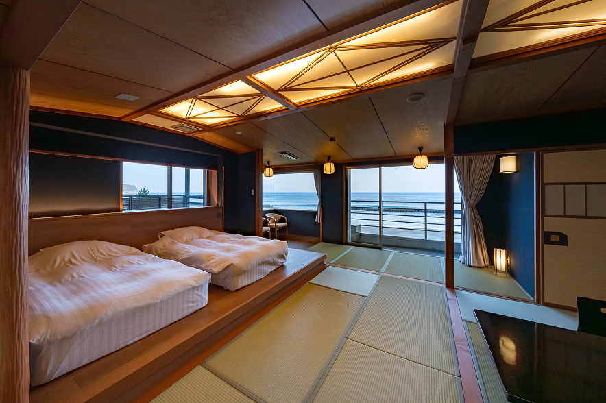 ベッドと畳のスペースが共存する広々とした客室。手前には、淡いピンクと白のストライプ柄のシーツがかけられた二つのベッドが、一段高くなった木製の床の上に並んでいる。ベッドの奥は畳敷きのエリアで、中央には光沢のある黒いテーブルと、背もたれのない座椅子が置かれている。