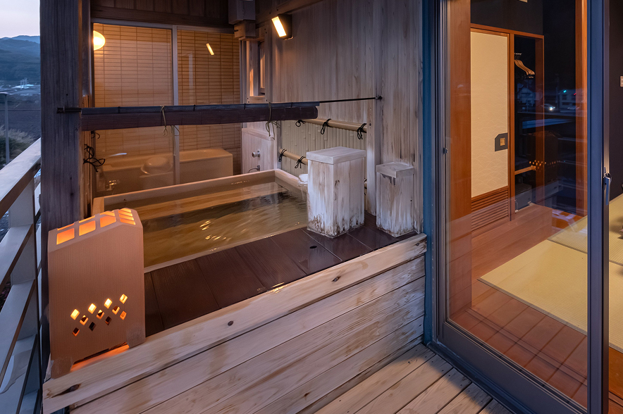 海辺の客室。手前には、白いストライプ柄のシーツがかけられた二つのシングルベッドが並んでいる。ベッドのヘッドボードは木製で、間接照明が温かい光を放っている。ベッドの奥は畳敷きのスペースになっており、中央には黒いローテーブルと座布団が置かれている。