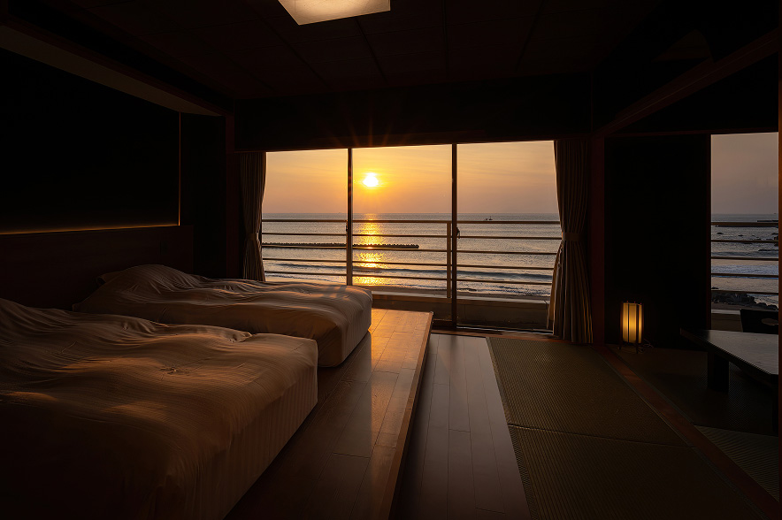 夕焼けに染まる海を望む旅館の一室。手前には、低い木製の台の上に二つのベッドが並び、夕日のオレンジ色の光を浴びて温かい色合いになっている。