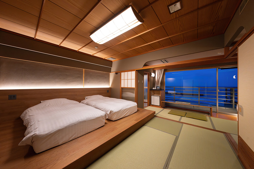 落ち着いた雰囲気の和モダンな客室。手前には、木製の低いベッドフレームの上に、ふっくらとした白いデュベスタイルの布団が二つ並んでいます。ヘッドボードの裏には間接照明が施され、温かい光が壁面を優しく照らしています。