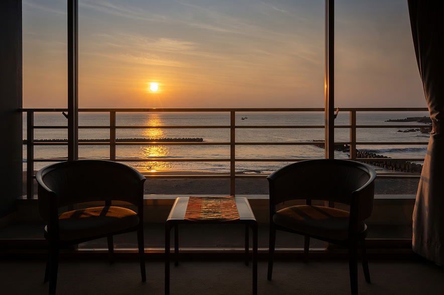 空が茜色に染まる、夕暮れのバルコニー。大きなガラス窓の外には、水平線に沈みゆく夕日が、海面にオレンジ色の光の道を創り出しています。
