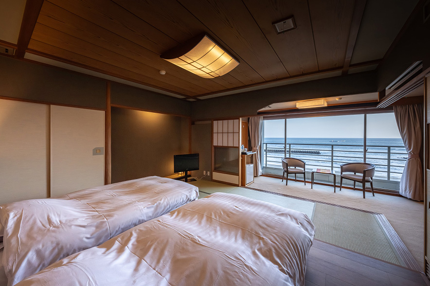 海辺の和モダンな客室。手前には、木製の低いベッドフレームの上に、清潔感のある白いストライプ柄の布団が二つ並んでいます。ヘッドボードの裏から漏れる柔らかな間接照明が、温かい雰囲気を醸し出しています。