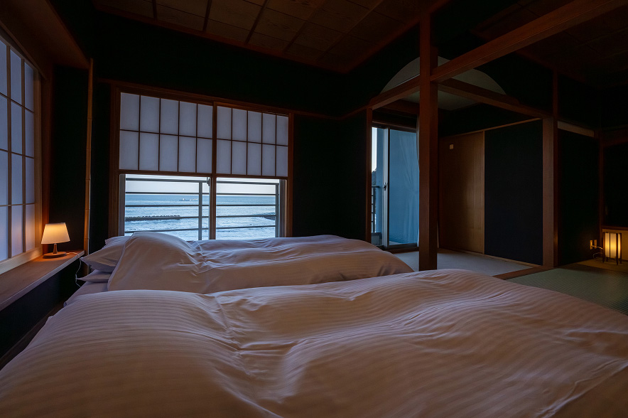 落ち着いた雰囲気の寝室。手前には、白いストライプ柄のシーツがかけられた二つのベッドが並んでいます。低い位置にあるベッドサイドランプが、温かいオレンジ色の光を静かに灯しています。