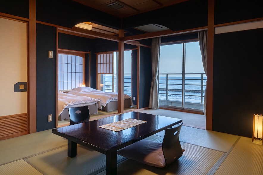 静かな朝の光が差し込む、海辺の客室。手前は畳敷きの居間スペースで、中央には光沢のある黒いローテーブルがあり、ベージュ色の和柄のランチョンマットが置かれています。