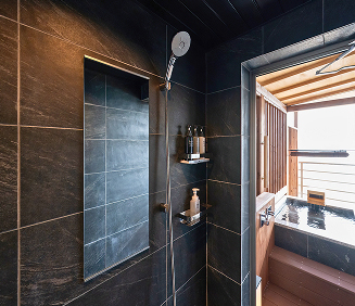 静かで穏やかな雰囲気の屋内露天風呂。濃い藍色の夕暮れの空が、木製の格子窓と小さなバルコニーへ続く開け放たれた戸口を通して見える。