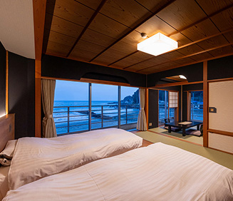 夕暮れ時の旅館の一室と露天風呂がガラス戸越しに見える風景。手前の露天風呂スペースは、木製の床と壁で囲まれており、湯船には温かい湯が張られている。