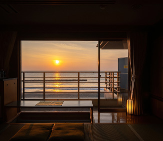 水平線から昇る朝日が、静かな部屋をオレンジ色に染める瞬間。大きなガラス窓の外には、穏やかな海が広がり、太陽の光が水面に一条の輝きを描いています。